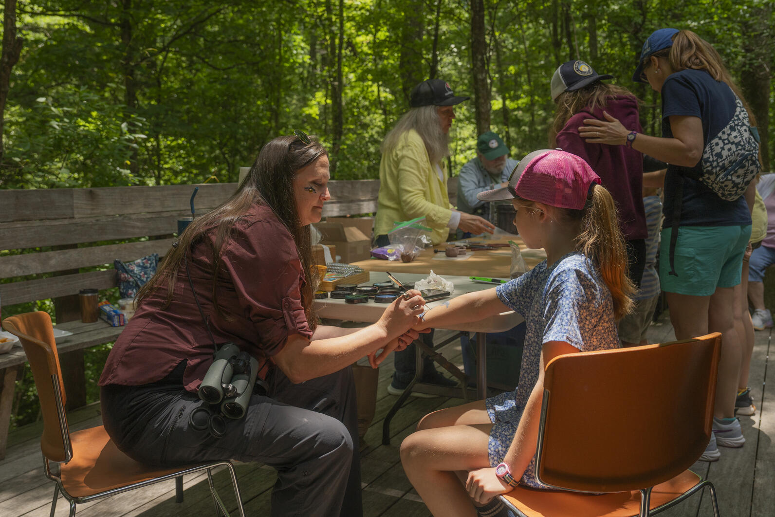 An Audubon staff person sits at a table on the backporch of the visitor center and paints on a girl's arm. Behind them two volunteers share nature objects with guests.