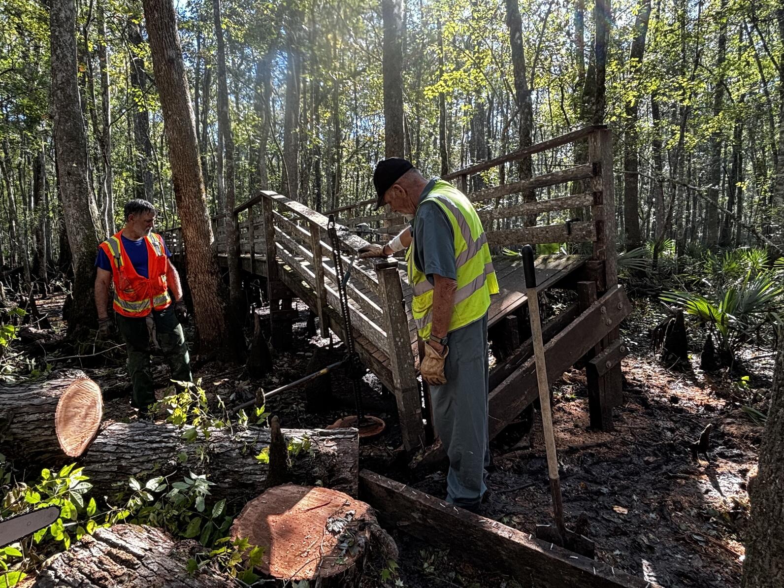 Mark Mussleman stands to the left of a metal jack position against a leaning section of boardwalk, in front of the section stands Rick Armstrong. At their feet are cut pieces of the sweetgum tree that crushed the boardwalk.