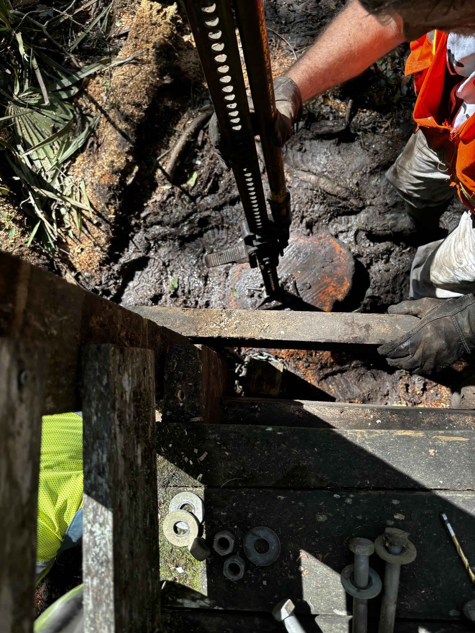 A metal jack is standing atop a wooden slice of a sweetgum tree, its upper section fit under the boardwalk to help lift it up when cranked.