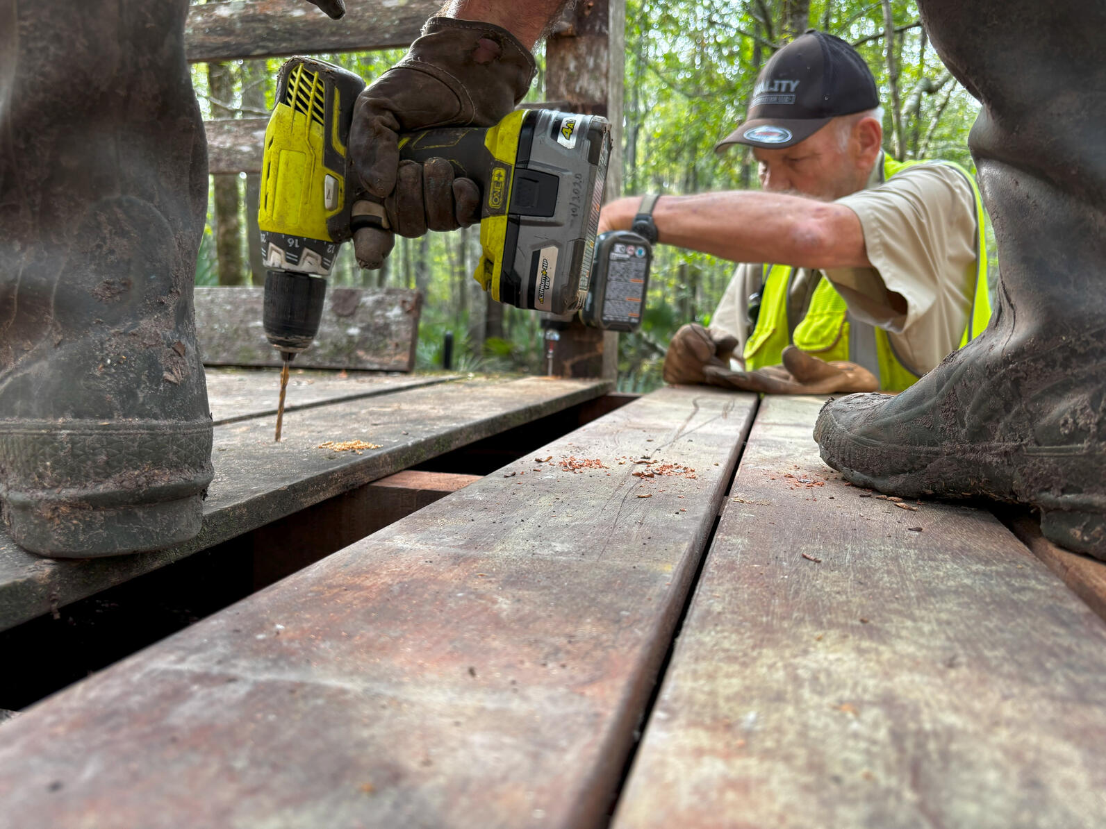 Mark stands on the boardwalk, using an electric drill on a deckboard to make holes for center screws of the walkway. Behind him on the ground stands Rick, who is leaning over the boardwalk to drill in holes for the edge screws.