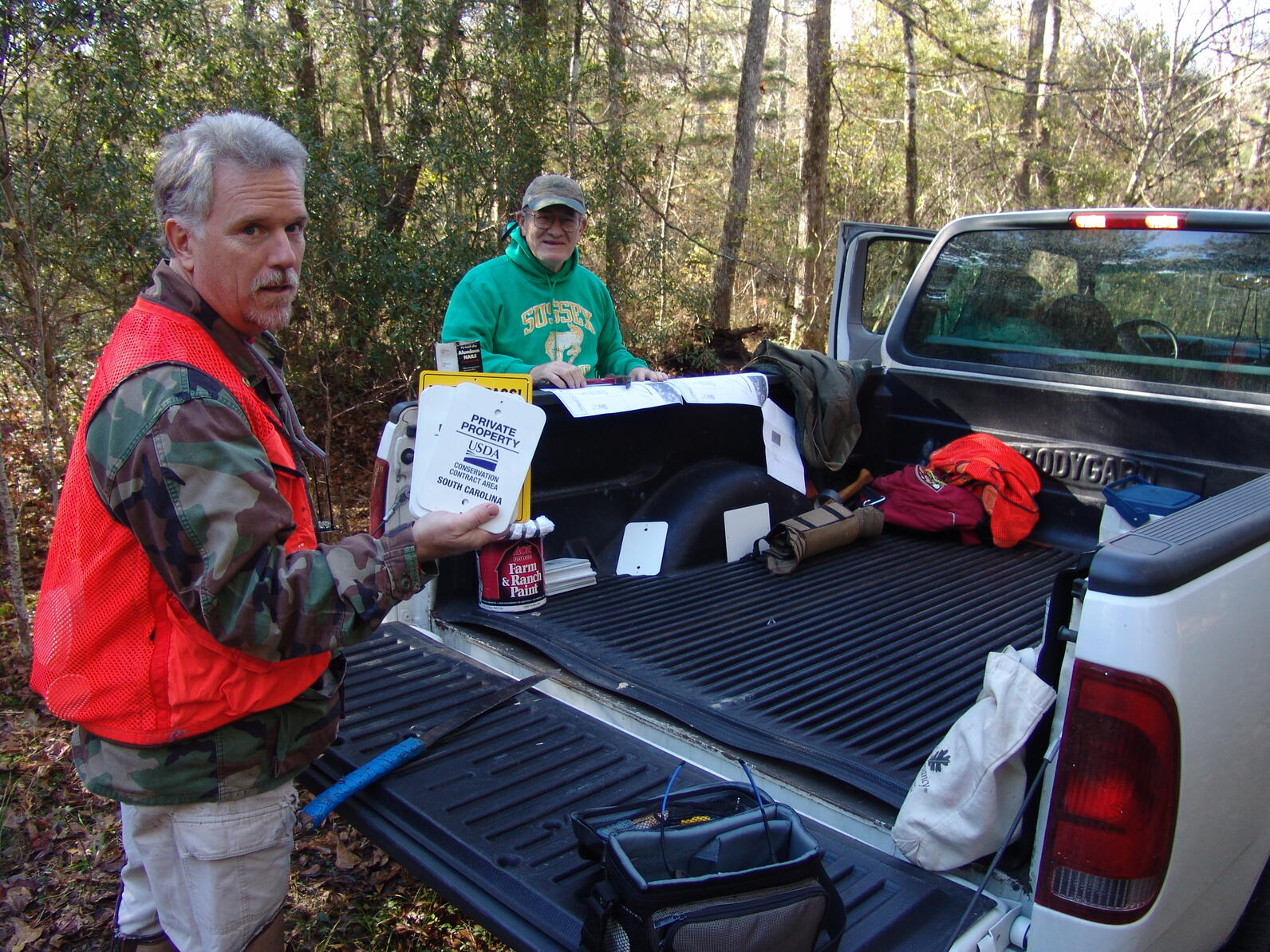 A man looks back at us and is holding signs put on the boundary of Beidler Forest's property to let others know who owns it. He is in front of a truck that has signs and tools in the back and resting against the truck is another man.