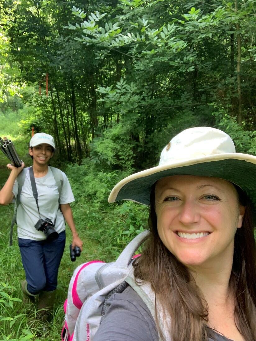 Two women are walking in dense forest edge with optical equipment and backpacks taking a selfie