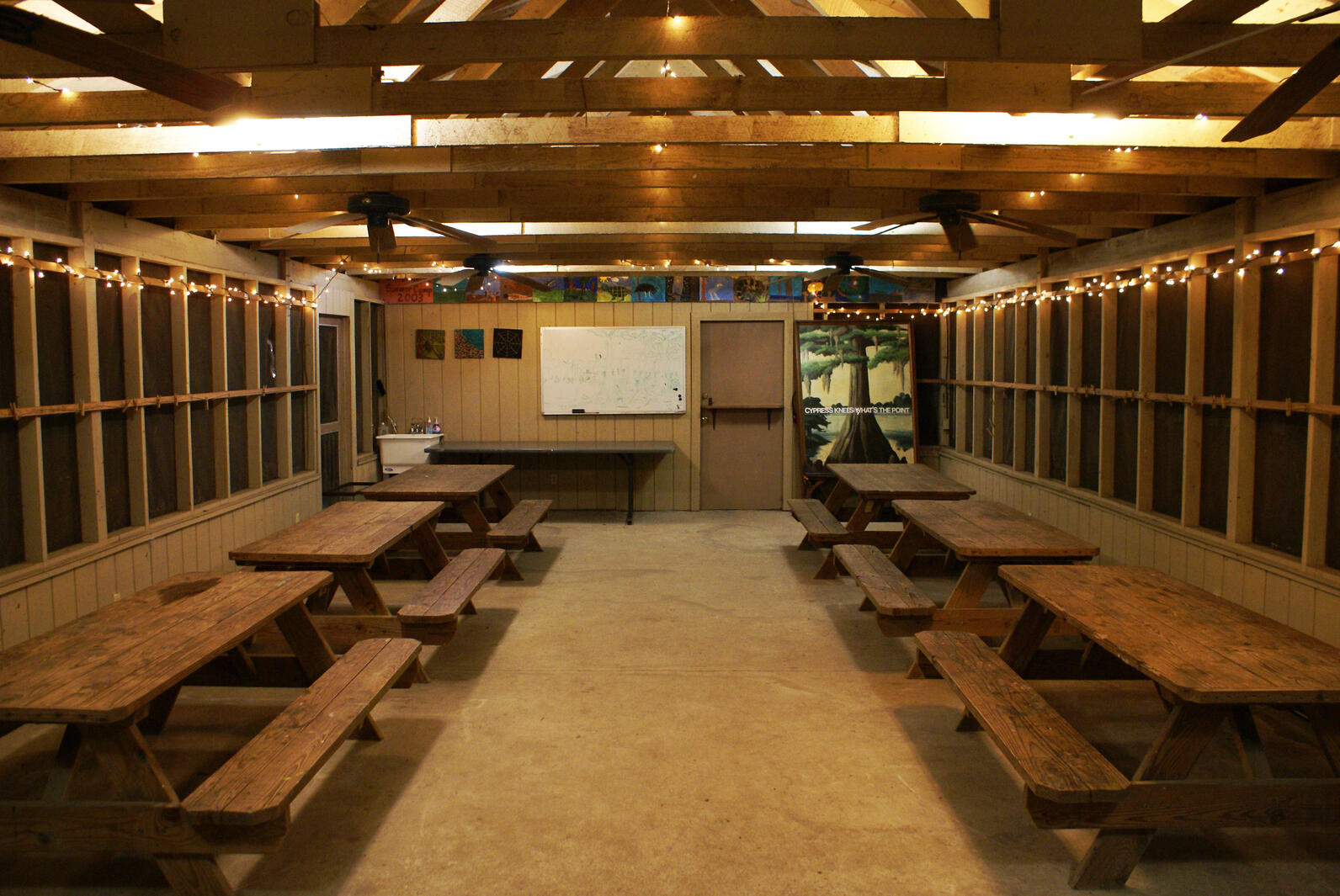 A view from inside Beidler's outdoor classroom. The screened-in walls are framed by rows of tables, three on either side. In the back of the room is another table, a sink, and a message board.