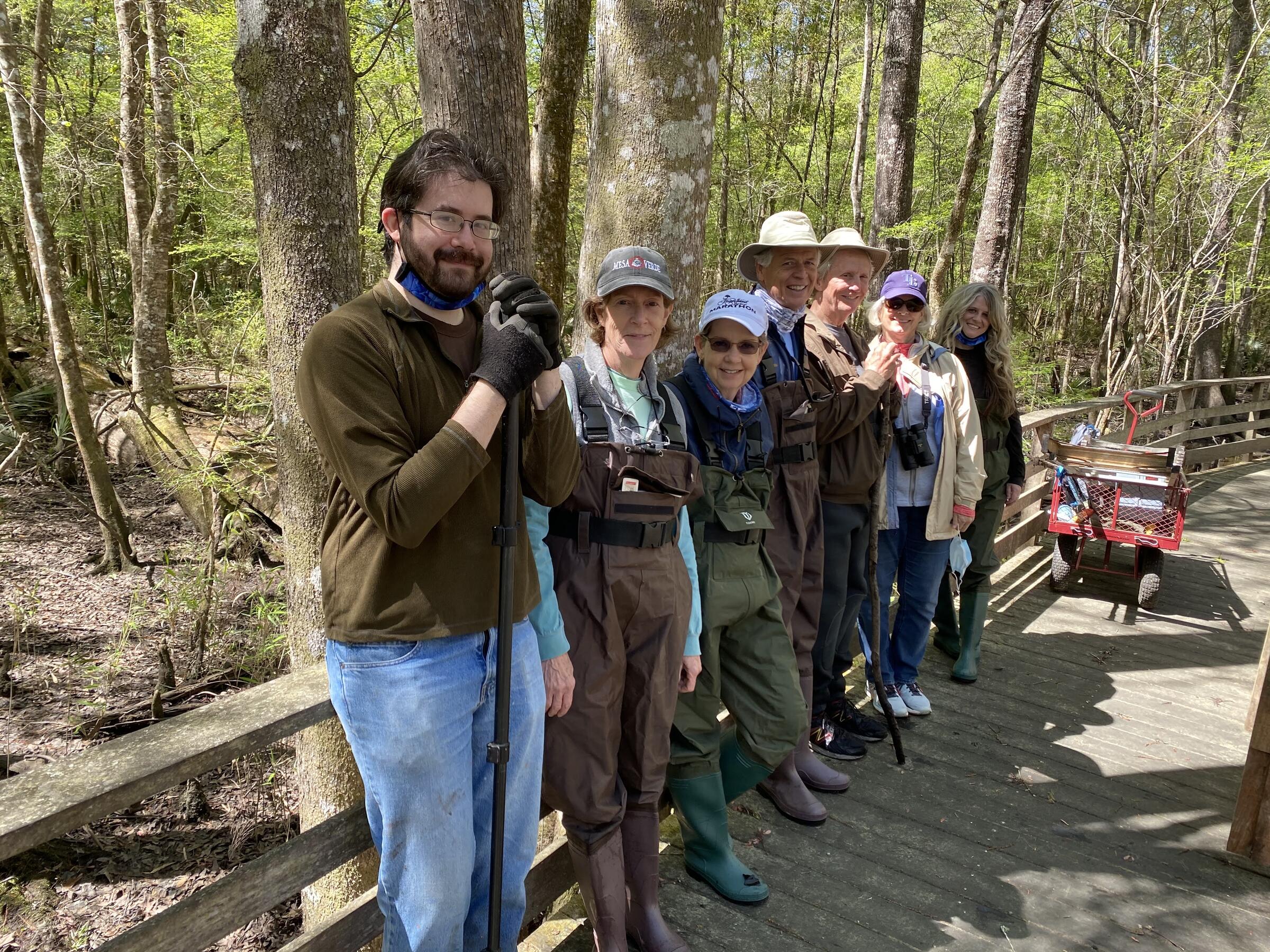 Seven people are lined up against the side of a boardwalk, some wearing waders and others wearing jeans. A red wagon with a ladder is in the distance and lush green forest behind the line of volunteers. 