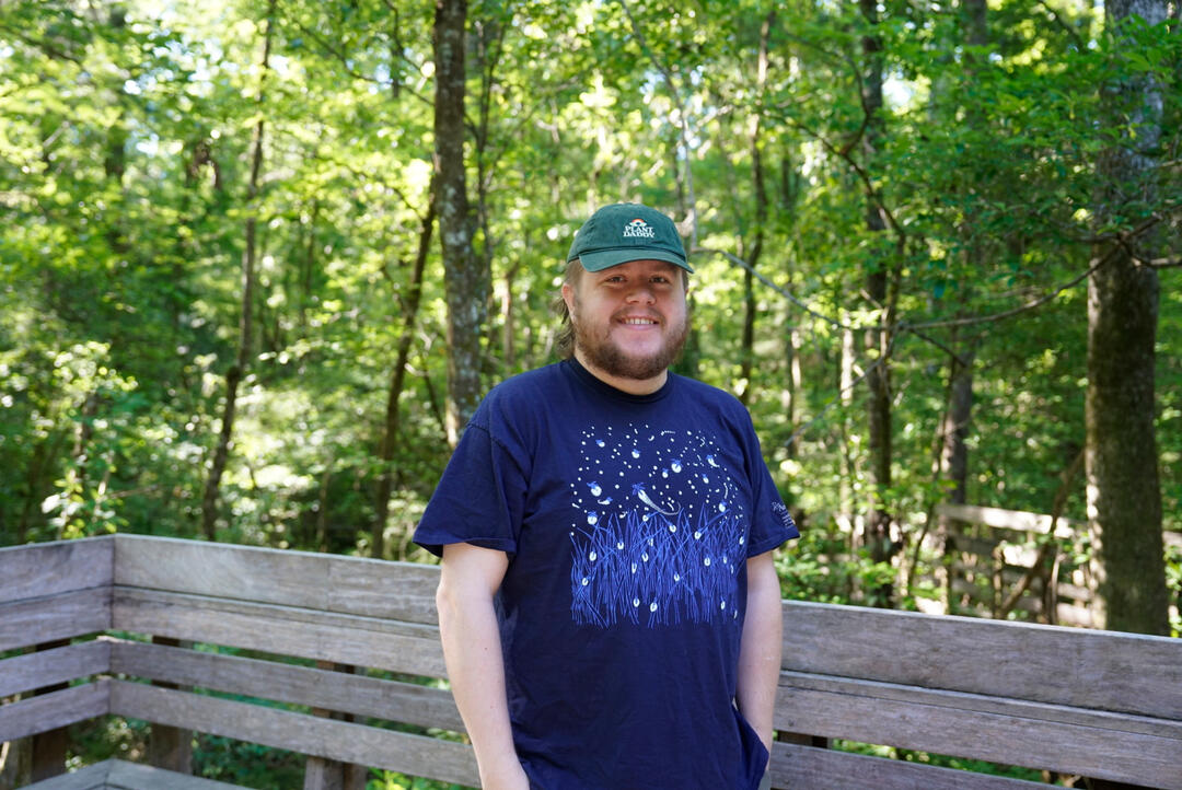Ryan Watson stands on the back porch of the visitor center smiling. He's wearing a firefly shirt and a ballcap with an owl on it.